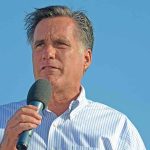 A man speaking into a microphone at an outdoor event against a blue sky