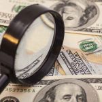 A magnifying glass resting on a pile of US dollar bills