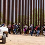 People walking beside tall fence and border patrol vehicle.