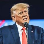 Trump Demands Clinton Probe—Epstein Emails Explode Man in suit speaking at a microphone.