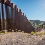 Trump Deploys Troops—Mexico Border Erupts Tall metal border wall with rural landscape