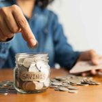 Person placing a coin into a savings jar labeled 'SAVINGS'