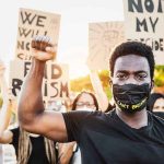 BLM Leader BUSTED — 25 Federal Fraud Charges Protester raises fist, wears mask, signs in background.