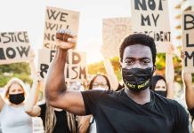 BLM Leader BUSTED — 25 Federal Fraud Charges Protester raises fist, wears mask, signs in background.