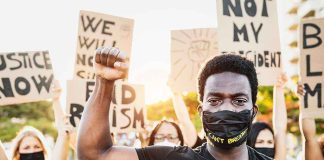 BLM Leader BUSTED — 25 Federal Fraud Charges Protester raises fist, wears mask, signs in background.