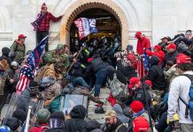 Pardoned J6 Rioters Launch REVENGE Campaign Crowd storming a building entrance with flags and signs.