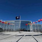 Trump’s F-35s Crush Russian Threats Flags outside NATO headquarters building under clear blue sky.