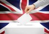 UK Political Elites in FULL Panic as New MAGA-Style Party Rises A hand placing a ballot into a white ballot box with the British flag in the background