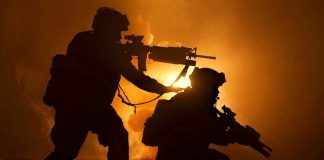 Two soldiers with weapons silhouetted against fiery background.