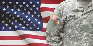 Soldier in camouflage uniform standing before American flag.
