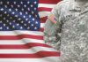 Retired Military Should Watching Closely Soldier in camouflage uniform standing before American flag.