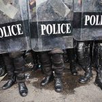 Police officers in riot gear standing behind shields marked POLICE