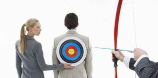 A woman places a target on a mans back while another man aims a bow and arrow at it