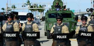 Police officers in riot gear with armored vehicle behind.