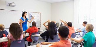 Beyond BELIEF — REMOVABLE Genital DOLLS APPROVED! Teacher in a blue dress instructing students in a classroom with hands raised
