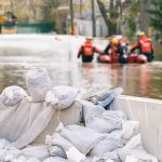 Deadly FLOODS Expose WIDESPREAD System Failures Sandbags stacked near a flooded street with rescue workers in a boat