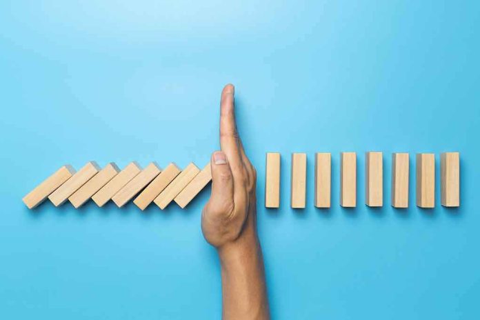 shutterstock_1891404868 Hand stopping falling wooden dominoes on blue background