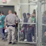 FBI Knocks After Chilling ICE Threat People standing inside chain-link holding area with an officer nearby