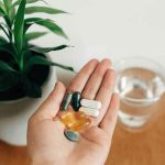 Hand holding assorted pills next to a plant and a glass of water