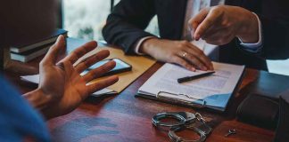 Two people discuss documents at a desk with handcuffs in view