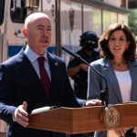 Alejandro Mayorkas speaks at a podium with Kathy Hochul standing beside him outdoors