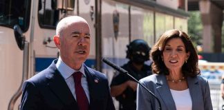 Alejandro Mayorkas speaks at a podium with Kathy Hochul standing beside him outdoors