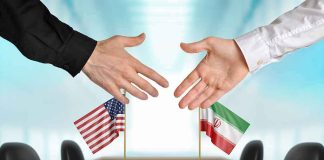 Two hands reaching to shake above US and Iran flags on a conference table