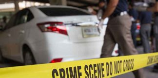 Yellow crime scene tape in front of a white car with investigators nearby