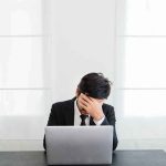 man in suit covers face while sitting at laptop