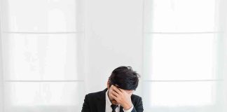 man in suit covers face while sitting at laptop