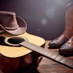 Acoustic guitar with cowboy hat and leather boots on wooden table