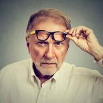 Older man lifting eyeglasses while looking intently at camera